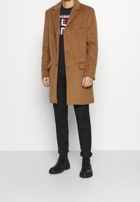 Abrigo de lana marrón con un solo botón y solapas de muesca, combinado con un suéter negro y pantalones de cuadros, llevado con botas chelsea negras.