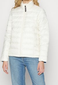 Biała pikowana kurtka z wysokim kołnierzem, zamek z przodu, dyskretna naszywka z logo na rękawie i teksturowany wzór pikowania. Noszona z niebieskimi dżinsami.