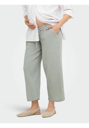 Personne enceinte portant un pantalon ample gris clair, une chemise blanche relevée pour montrer le ventre, et des chaussures plates beiges tissées.