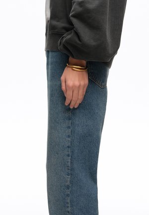 Osoba ubrana w niebieskie dżinsy i ciemną koszulę z długim rękawem, na lewej ręce nosząca złotą skręconą bransoletkę.