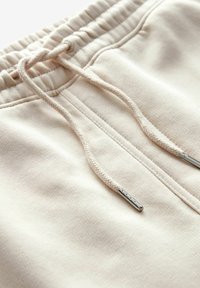 Pantalons de survêtement beige clair en tissu texturé, avec une taille élastique et un cordon de serrage tressé avec des embouts métalliques. Design simple et fonctionnel.