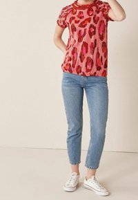 Person iført kortærmet top med rødt og pink leopardmønster, lyseblå cropped jeans og hvide sneakers, stående mod en ensfarvet baggrund.
