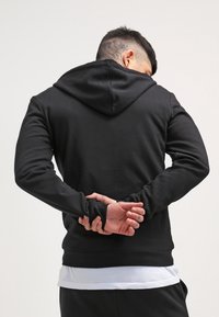 Sudadera negra con vista trasera, que presenta una capucha considerable, mangas largas y un diseño limpio y sencillo. La tela parece suave y lisa.