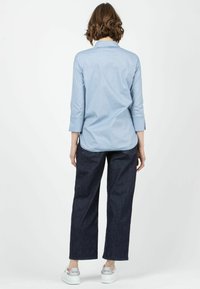 Camisa de botón azul claro con mangas tres cuartos, combinada con pantalones de mezclilla anchos de color oscuro y zapatillas blancas. Diseño minimalista.