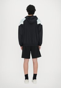 Jachetă neagră cu un design ușor albastru pe umăr, cu glugă și talie elastică. Asortată cu pantaloni scurți negri și adidași albi, văzută din spate.