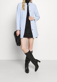Donna che indossa un cappotto azzurro chiaro con bottoni, dolcevita nero e gonna, stivali neri fino al ginocchio, mentre tiene una grande pochette nera con texture.
