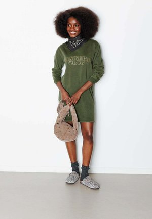 Femme à la coiffure afro en robe verte à manches longues tenant un sac beige, portant des chaussettes grises et des chaussures à enfiler imprimé léopard, souriante.