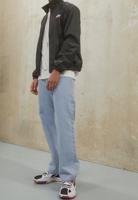 Svart nylonjacka med elastiska mudd, vit logga. Ljusblå baggy-jeans, svarta och vita sneakers med färgade detaljer och snören.