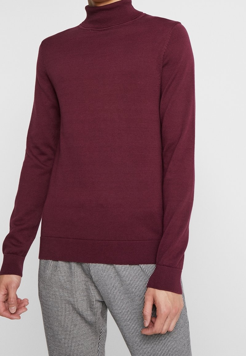 Maglione a dolcevita bordeaux realizzato in morbido tessuto a maglia. Presenta maniche lunghe e orlo a coste. Indossato con pantaloni a scacchi.