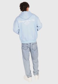 Mann steht, trägt einen hellblauen Hoodie mit weißen Text auf dem Rücken, hellblaue Jeans und weiße Sneaker, mit dem Rücken zur Kamera.