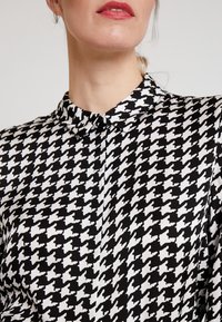 Sort og hvid skjorte med knappesystem præget af et houndstooth-mønster. Stoffet virker glat med en klassisk krave og knaplukning.