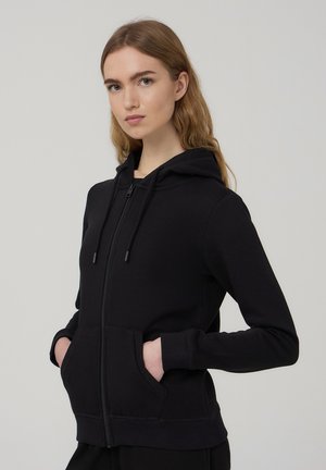 Felpa nera con zip, dotata di tasca a marsupio frontale, cappuccio con coulisse, polsini a costine e un tessuto morbido e texturizzato.