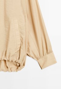 Ljusbeige tröja med en texturerad yta, med elastiska manschetter och en sidficka, designad med en avslappnad passform.