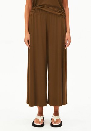 Person, die lockere braune Hosen mit weitem Bein und weiße Sandalen mit schwarzen Sohlen trägt und vor einem weißen Hintergrund steht.
