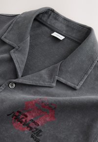 Camicia grigio scuro con colletto, bottone e motivo ricamato rosso e nero sul petto, stesa piatta su una superficie chiara.