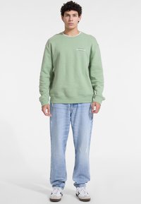 Svetlo zeleni pulover z rebrastimi manšeti in robovi, ki ima majhen logotip. Kombiniran s svetlo modrimi unikatnimi jeans hlačami in belimi supergami.