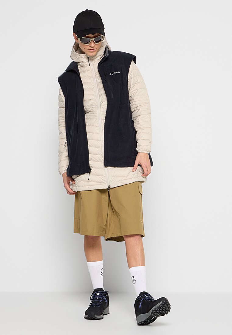 Persoon met een zwarte pet, zonnebril, een lichtbeige pufferjack met een zwart mouwloos vest, kaki shorts, witte sokken en zwarte wandelschoenen.