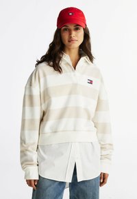 Beige en witte gestreepte pullover met een kraag en knopenopening, gecombineerd met een wit shirt en blauwe jeans. Rode pet met logo.