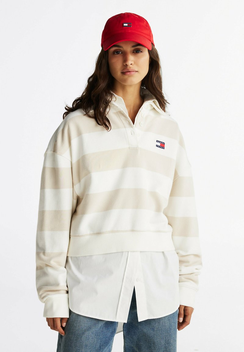 Beige en witte gestreepte pullover met een kraag en knopenopening, gecombineerd met een wit shirt en blauwe jeans. Rode pet met logo.