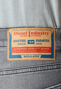 Brun läderetikett med blå och röd tryck, som står "Diesel Industry." Texten nämner "Överlägsen Kvalitet Jeans" och "Reglerad," fäst vid grå denim.
