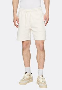 Kermanväriset puuvillahousut, joissa on joustava vyötärönauha ja kiristysnyöri, ja joissa on yksinkertainen logo lahkeessa. Yhdistetty vaalean värisiin kenkiin ja sukkiin.