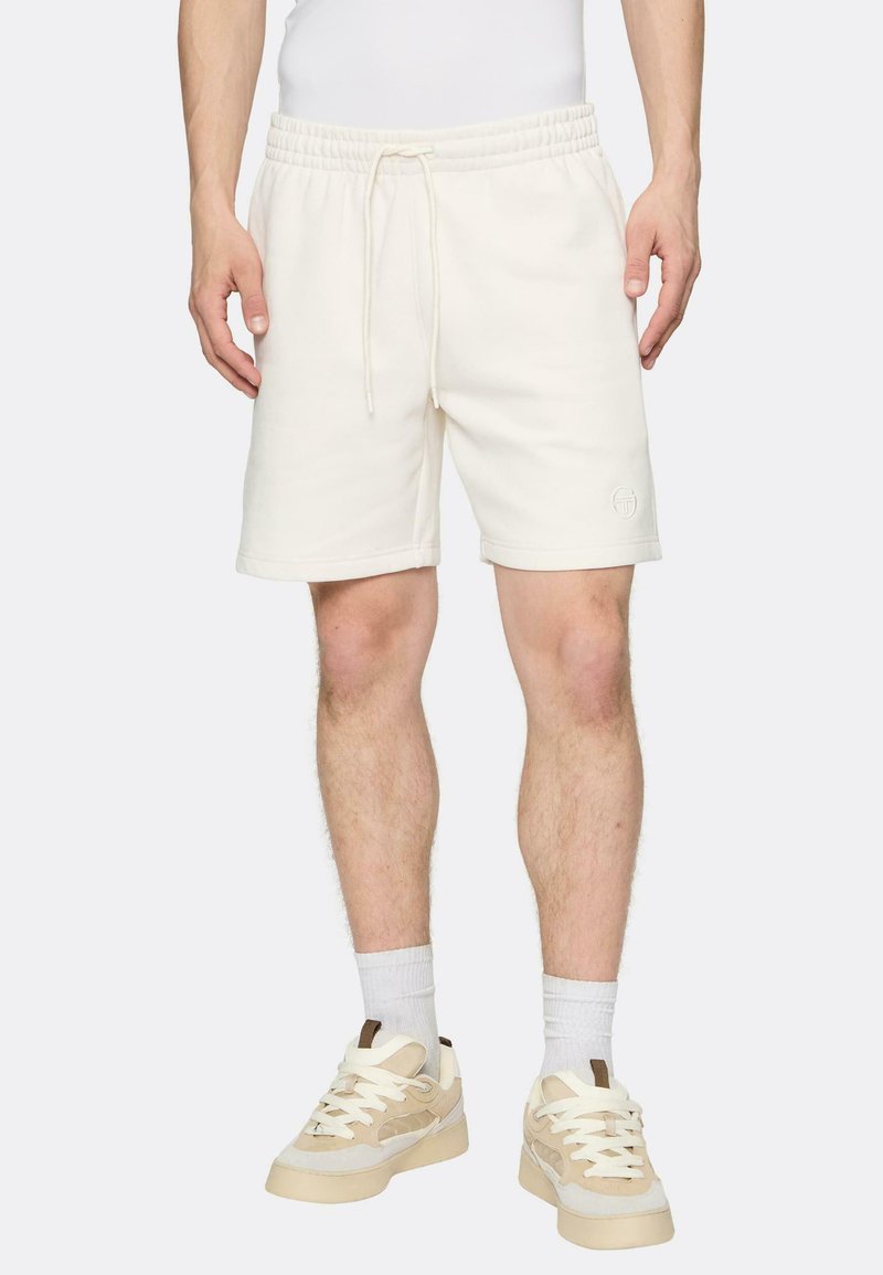 Kermanväriset puuvillahousut, joissa on joustava vyötärönauha ja kiristysnyöri, ja joissa on yksinkertainen logo lahkeessa. Yhdistetty vaalean värisiin kenkiin ja sukkiin.