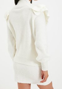 Weißer gestrickter Pullover mit Rüschenakzenten an den Schultern und gerippter Textur. Mit langen Ärmeln und einer figurnahen Silhouette. Kombiniert mit einem passenden Miniröcke.