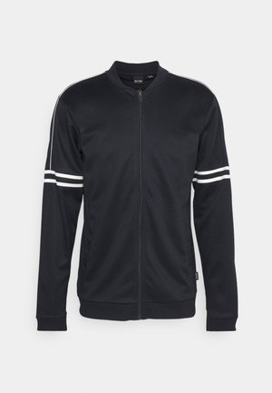 Veste noire à zip avec doubles bandes blanches autour des bras supérieurs et un fin liseré blanc le long des épaules et des manches.