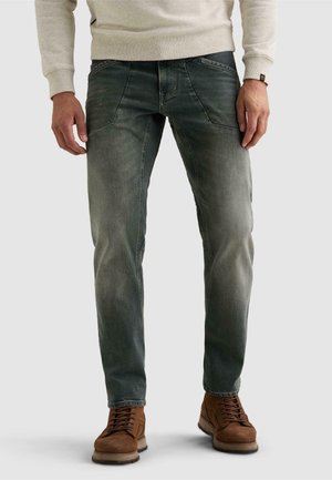 Homme portant un jean noir délavé, des bottes marron à lacets et une chemise à manches longues beige clair, debout devant un fond uni.
