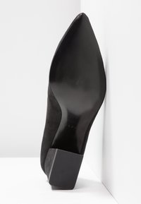 Chaussure à talon haut noire à bout pointu avec talon bloc texturé, semelle orientée vers l'avant sur un fond blanc en coin.