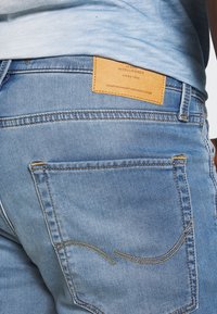 Blue denim jeans with a light wash, featuring yellow stitching and a brown leather patch on the back with brand details.