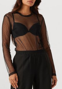 Top lungo a maniche trasparenti nere in mesh con texture a pois, abbinato a un reggiseno nero. La modella indossa pantaloni neri con vita elastica.