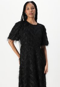 Robe noire texturée avec manches courtes bouffantes, motif zigzag et détails frangés. Tissu lisse avec des accents brillants partout.