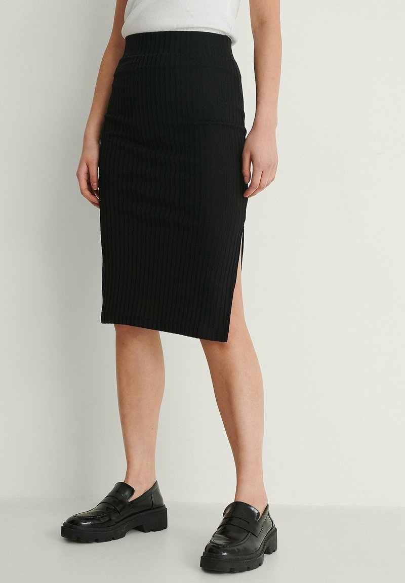 Jupe midi côtelée noire à taille haute, avec des fentes latérales. Associée à des mocassins noirs épais. Texture lisse.