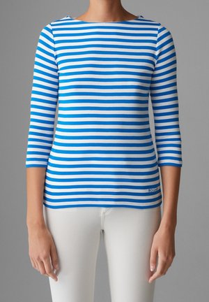Femme portant un haut à manches trois-quarts rayé horizontalement bleu et blanc avec un pantalon blanc sur un fond gris uni.