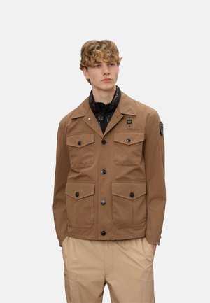 Giovane uomo con capelli ricci che indossa una giacca marrone con bottoni e molte tasche e pantaloni beige, in piedi con le mani nelle tasche.