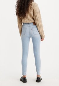 Jeans de corte slim en azul claro con una altura alta; cuentan con bolsillos traseros y una etiqueta de cuero marrón en la cintura; combinados con un suéter texturizado en beige.