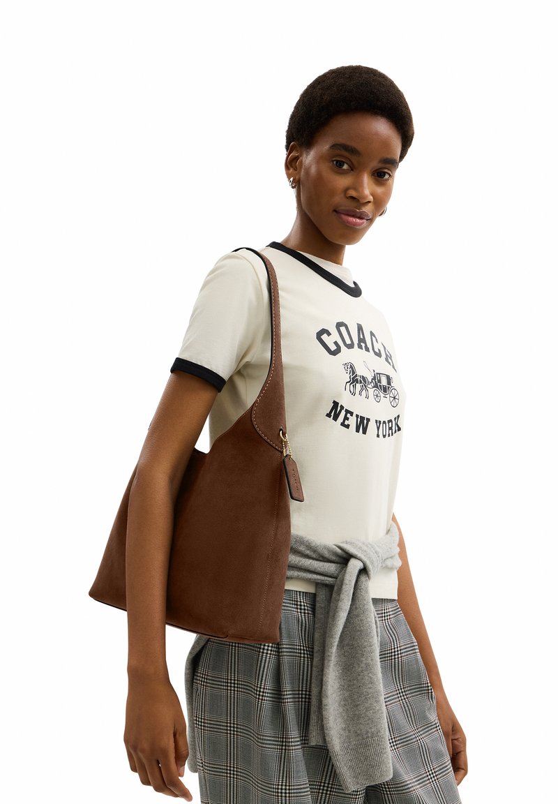 Femme portant un t-shirt blanc Coach New York, un pull gris noué à la taille, un pantalon à carreaux et portant un grand sac à bandoulière en cuir marron.