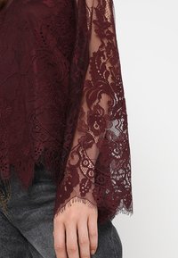 Burgunderrød bluse i blonde med indviklede blomsterdesigns, gennemsigtig tekstur, klokkeærmer og bølgekant. Sammen med mørke denimjeans.