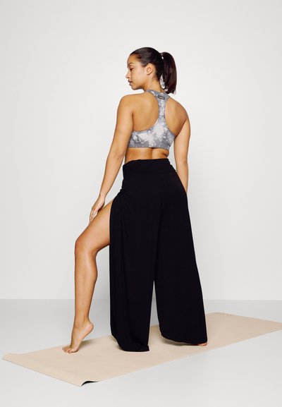 Sutiã desportivo cinza com efeito tie-dye e design de costas nadador, combinado com calças largas pretas com uma fenda alta, usadas sobre um tapete de yoga.