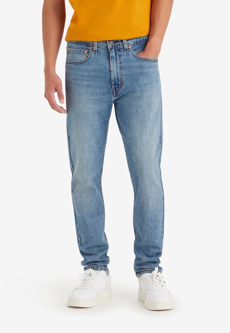 Homme portant un jean denim bleu clair, chemise orange, baskets blanches, avec une main dans la poche et l'autre détendue le long du corps.