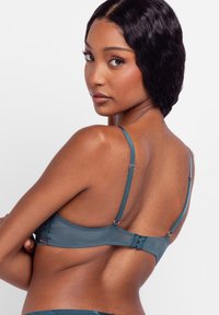 Woman with smooth skin and wavy dark hair wearing teal adjustable-strap bra, looking over shoulder against white background.