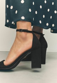 Sandales à talons hauts en suède noir avec un talon bloc, bout ouvert, et une sangle de cheville dotée d'une boucle réglable. Ourlet de robe à pois en arrière-plan.