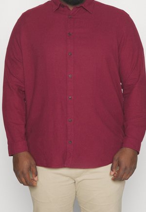 Homme portant une chemise bordeaux à manches longues boutonnée et un pantalon beige, les mains détendues le long du corps, sur un fond uni.