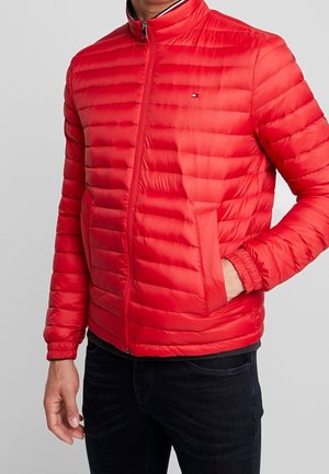 Mann trägt eine leuchtend rote Steppjacke mit horizontaler Steppung, Frontreißverschluss und Seitentaschen, kombiniert mit dunkler Hose.