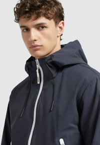 Veste coupe-vent marine avec zipper blanc, capuche ajustable et cordons. Présente une texture lisse et un design minimaliste.