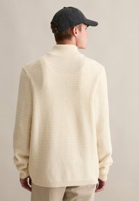 Cremefarbener, strukturierter Strickpullover mit einem hohen Kragen und gerippten Bündchen, kombiniert mit einer dunklen Mütze. Die Rückseite zeigt einen lockeren Schnitt und ein gewebtes Muster.