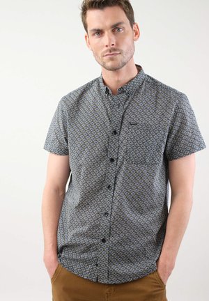 Chemise à manches courtes à boutons en tissu sombre, ornée d'un motif géométrique de diamants bleus et beiges, avec une poche poitrine et un col structuré.