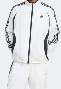 Hombre con chándal blanco de Adidas con rayas negras a lo largo de las mangas y los costados, manos en los bolsillos, chaqueta con cremallera, logo visible en el pecho y los pantalones.