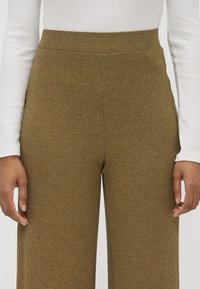 Olivgrön ribbad byxor med vida ben och hög midja, med ett strukturerat tyg. En vit långärmad topp bärs under.